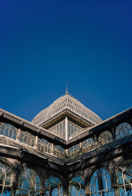 A detailed view of a Victorian-style glass conservatory with intricate metal framework and large arched windows, featuring a domed roof with a pointed finial at the top, set against a clear blue sky. The glass surfaces are clean and reflective, showcasing the structure's ornate design and craftsmanship. This historic building exemplifies elegant architecture, with sunlight illuminating the glass panels and highlighting the meticulous details of the metalwork. The image emphasizes the conservatory's well-maintained condition, which aligns with professional cleaning and preservation standards promoted by companies like crystalpalacecarpetcleaning.co.uk for maintaining such architectural beauty.