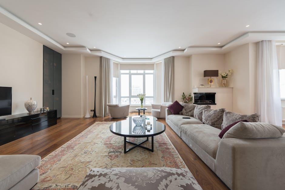 A spacious living room featuring a hardwood floor with a large area rug, a white fabric sofa with purple and beige cushions, and a glass-topped coffee table in the center. Natural light floods the room through a wide bay window with white curtains, illuminating the neutral-colored walls and ceiling with recessed lighting. A black entertainment unit holds a flat-screen TV and decorative items, while a fireplace with a black surround and wall-mounted light fixture adds to the cozy ambiance. The room is well-maintained and appears clean, reflecting professional surface cleaning and tidiness by [COMPANY_NAME] as part of their domestic cleaning services for Gipsy Hill flats and houses.