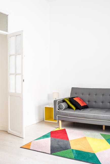 A bright, minimally decorated living room featuring a grey upholstered sofa with tufted backrest and a variety of colorful cushions in green, black, red, and grey. The room has white walls and light wooden flooring, with a multicolored geometric-patterned rug positioned in front of the sofa. To the left, there is a partially open white wooden door with glass panels, allowing natural light to illuminate the space. A small yellow side table with a white lamp is placed next to the sofa. The overall scene depicts a clean, tidy, and well-maintained domestic setting with an emphasis on surface cleaning and home hygiene, illustrating a typical area that professional cleaning services like those offered by crystalpalacecarpetcleaning.co.uk might service for deep cleaning or sanitisation.