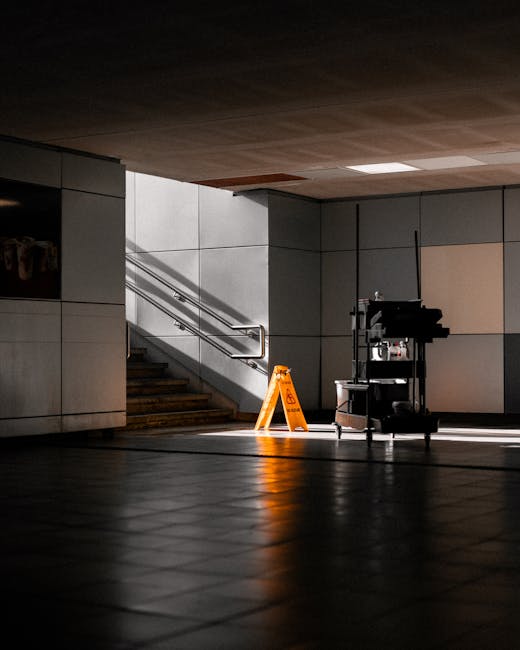 A dimly lit underground or subway station entrance with a tiled floor, grey concrete walls, and a staircase with metal handrails leading upwards. In the center, there is a small cleaning cart equipped with bottles and cleaning supplies, along with a yellow caution sign indicating wet floor, suggesting recent or ongoing cleaning. The lighting creates shadows on the floor and walls, emphasizing the cleanliness and maintained condition of the area. This scene likely relates to professional surface cleaning or sanitisation services offered by crystalpalacecarpetcleaning.co.uk for commercial spaces such as transit hubs.