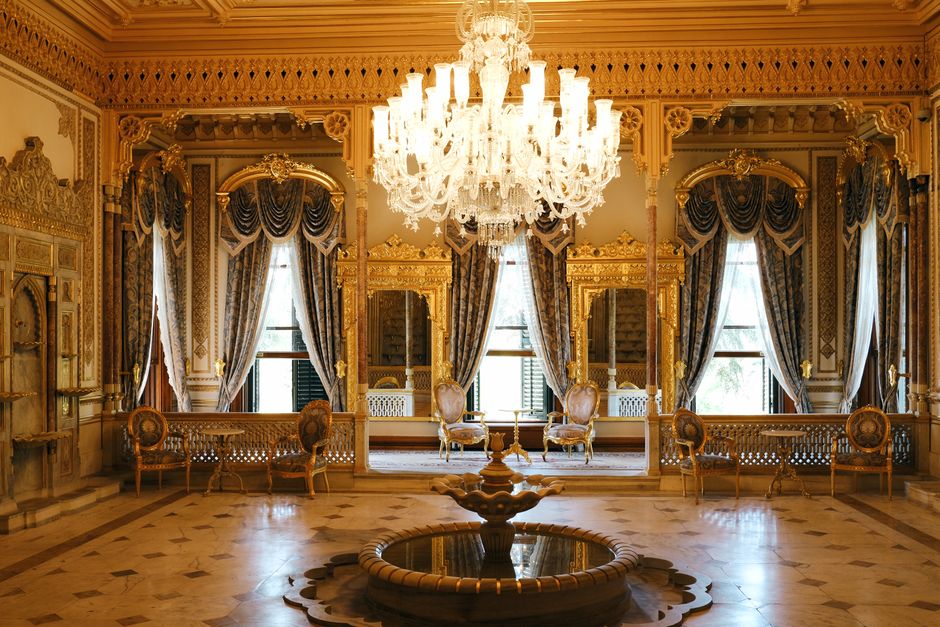 An opulent, luxurious room featuring ornate, gold-accented moldings and detailed wall paneling, illuminated by a large, intricate crystal chandelier hanging from a decorated ceiling. There are tall, elegant windows with heavy, dark blue drapes and valances, allowing natural light to filter into the space. The polished wooden floor displays a herringbone pattern and reflects the ambient light, enhancing the room's grandeur. Opposite the windows, a central decorative water fountain is situated on the floor, surrounded by a small circular platform. Along the walls, antique-style chairs with gold frames and upholstered seats are neatly arranged, some positioned near the windows. The room appears highly clean and well-maintained, exemplifying careful surface cleaning and decorative item dusting, consistent with the services provided by crystalpalacecarpetcleaning.co.uk for upholstery cleaning and deep cleaning of luxurious spaces.