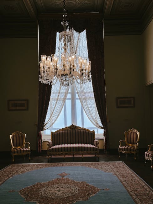 A luxurious sitting room featuring a large, elegant crystal chandelier hanging from a dark wooden ceiling panel, casting a soft glow over the space. Beneath the chandelier, there is a vintage-style upholstered sofa with vertical stripes in shades of pink and gold, flanked by matching armchairs on either side. The room has tall, ornate windows draped with white lace curtains and dark, heavy drapes that are pulled back, allowing natural light to filter in. The floor is covered by a patterned area rug with intricate designs in muted tones. The walls are painted in a neutral color, with small decorative frames hanging symmetrically on each side of the window. The overall appearance of the room is pristine, orderly, and well-maintained, reflecting a high standard of cleaning and care, which is associated with services offered by crystalpalacecarpetcleaning.co.uk for upholstery cleaning and deep cleaning applications in historic or elegant residential settings.